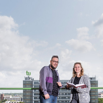 Zwei Studierende blicken in die Kamera, die Studentin (rechts) hält ein aufgeschlagenes Buch in der Hand, im Hintergrund ist der Mathetower in seiner vollen Länge zu sehen. 