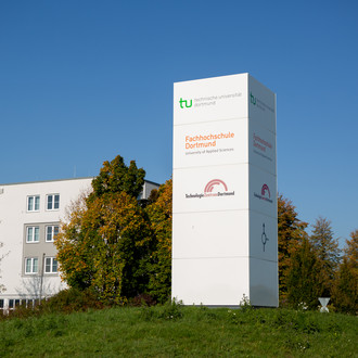 Hinweisschild auf einem Kreisverkehr, im Hintergrund ein Gebäude und Bäume.