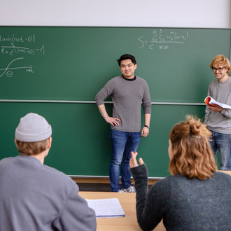 Zwei Studierende sitzen im Vordergrund im Seminarraum. Im Hintergrund stehen der Dozierende und ein Studierender an der Tafel