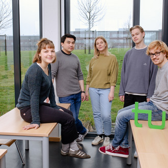 Fünf Studierende sitzen auf dem Tisch und stehen beisammen in einem Seminarraum.