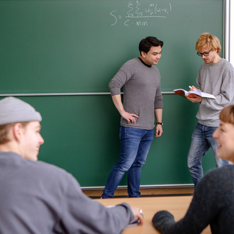 Zwei Studierende sitzen im Vordergrund im Seminarraum. Der Dozierende und ein Studierender stehen im Hintergrund an der Tafel.