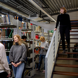 Zwei Studierende stehen neben Bücherregalen und unterhalten sich. Ein Studierender geht die Treppe herunter.