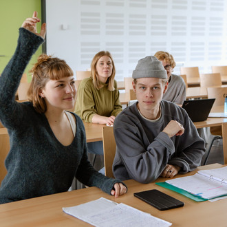 Studierende im Seminarraum sitzen an den Tischen. Eine Person meldet sich. 