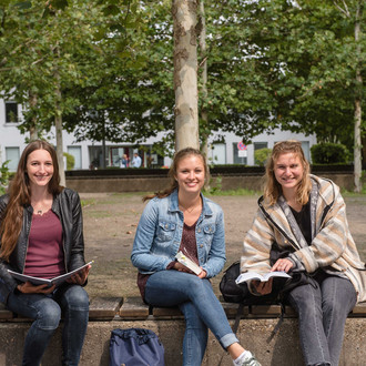 Drei Studentinnen sitzen auf einer Bank, halten Bücher in ihren Händen und lächeln.