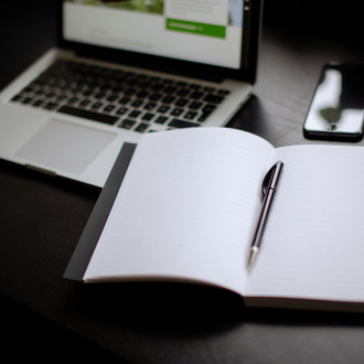 Eingeschalteter Laptop, Smartphone und Notizbuch mit Stift auf einem Tisch liegend.