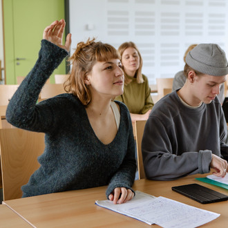 Studierende im Seminarraum sitzen an den Tischen. Eine Person meldet sich. 