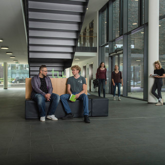 Zwei Studierende sitzen auf einer Bank und unterhalten sich. Andere laufen vorbei.