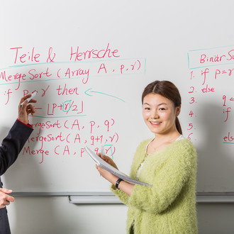 Zwei Personen stehen vor einem Whiteboard mit Formeln und Beschriftung. Eine Person hat einen Stift in der Hand, die andere einen Block.