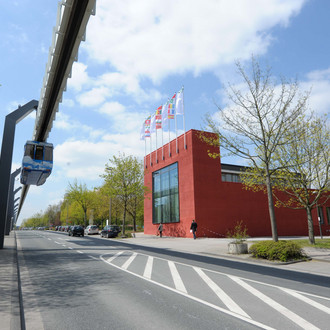 Rotes Gebäude umgeben von Bäumen mit blauem Himmel und wenigen Wolken. Links im Bild die H-Bahn.