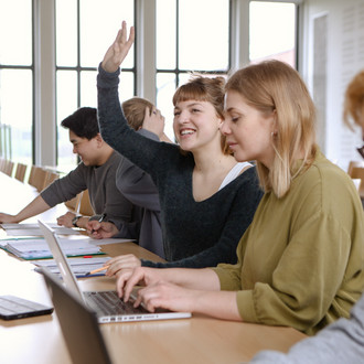 Fünf Studierende sitzen in einem Seminarraum an Tischen. Eine Studentin hebt ihre Hand.