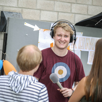 Ein Student mit Kopfhörern auf dem Kopf und Mikrofon in der Hand unterhält sich mit einem Studenten und einer Studentin, die an seinem Stand stehen.