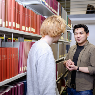 Zwei Studierende unterhalten sich zwischen Regalreihen in der Bibliothek.