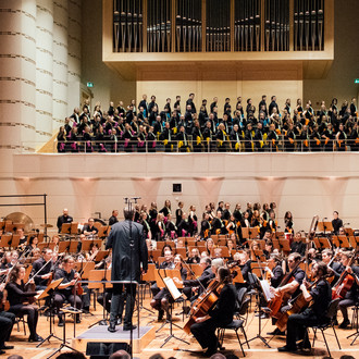 Ein Orchester spielt auf einer Bühne Musik. Über der Bühne steht auf einer Empore ein Chor.