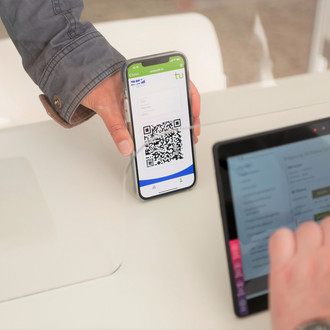 Eine Hand hält ein Smartphone mit einem QR-Code in der TU-App, der von einem Tablet gescannt wird.