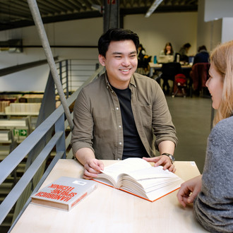 Zwei Studierende unterhalten sich an einem Tisch in der Bibliothek und lächeln sich an.