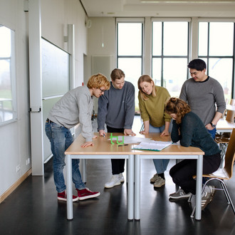 Fünf Studierende stehen im Seminarraum um einen Tisch und sehen auf ein Dokument