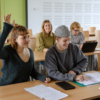 Studierende im Seminarraum sitzen an den Tischen. Eine Person meldet sich. 