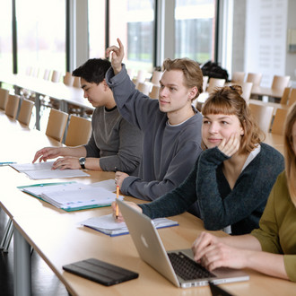 Studierende im Seminarraum sitzen in einer Tischreihe vor ihren Laptops. Ein Studierender meldet sich.