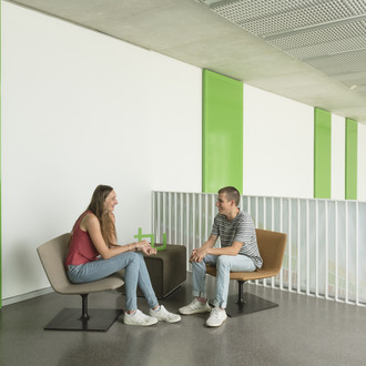 Zwei Studierende sitzen auf Stühlen an einem Tisch, auf dem das TU Dortmund Logo steht. Hinter ihnen befindet sich ein Handlauf und eine Treppe.