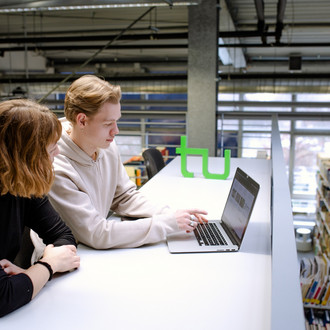 Zwei Studierende sitzen in der Bibliothek an einem langen Tisch und schauen gemeinsam auf einen Laptop.