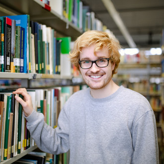 Student steht zwischen Bücherregalen.