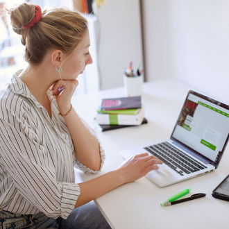 Am Schreibtisch sitzende Studentin tippt auf dem Laptop, daneben liegen Bücher und Stifte.