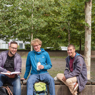 Drei Studenten sitzen vor grünen Bäumen auf einer Bank.