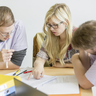 Drei Personen sitzen an einem Tisch und haben Unterlagen vor sich liegen. Die Person in der Mitte zeigt auf die Unterlagen und spricht.