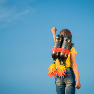 Man sieht eine junge Person mit gelbem T-Shirt, Jeans und Lederkappe von hinten vor einem blauen Himmel. Sie hat eine Attrappe eines Raketenrucksacks auf dem Rücken und streckt die linke Faust in die Höhe.