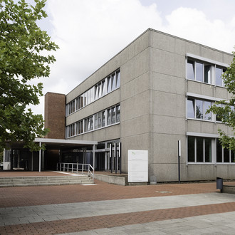 HGB I: In der Mitte ein graues Gebäude von der Seite fotografiert. Links und rechts Bäume. Rot-Graue Steine auf dem Boden.