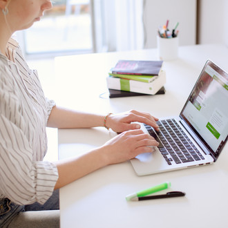 Am Schreibtisch sitzende Studentin tippt auf dem Laptop, daneben liegen Bücher und Stifte.