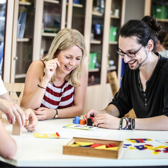 Zwei Personen sitzen an einem Tisch. Die Person rechts hat die Hände um bunte Bausteine auf dem Tisch gelegt. Beide gucken auf die Bausteine auf dem Tisch und lachen. Links auf der gegenüberliegenden Seite des Tisches sitzt eine weitere Person, die man von hinten sieht.