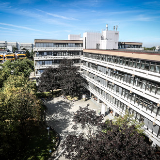 Emil-Figge-Straße 50: Ein langes Gebäude mit Fenstern, vor denen teilweise Rollos heruntergezogen sind, von oben fotografiert. Davor ein Platz mit Bäumen. Hinten links im Hintergund das gelb-weiße Mensa-Gebäude.