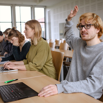 Studierende im Seminarraum sitzen in einer Tischreihe vor ihren Laptops.  Eine Person meldet sich.