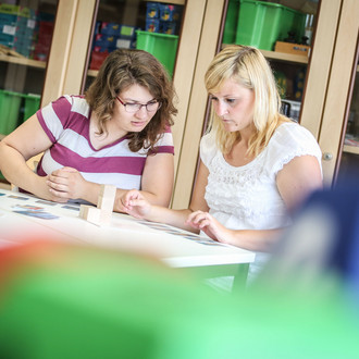 Zwei Personen sitzen an einem Tisch und beschäftigen sich konzentriert mit Bausteinen und Kärtchen, die auf dem Tisch liegen. Im Hintergrund steht ein Schrank voll mit Kisten, Scheren und anderen Materialien.