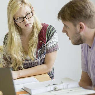 Nahaufnahme von zwei Personen, die an einem Tisch sitzen. Die Person rechts hat Unterlagen vor sich liegen und spricht. Die Person links hört der anderen Person konzentriert zu.