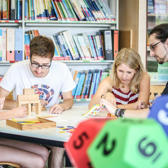 Vier Personen sitzen an einem Tisch und beschäftigen sich mit Bausteinen, die auf dem Tisch liegen. Im Vordergrund sieht man bunte vieleckige Würfel. Im Hintergund steht ein Regal voller Bücher und Ordner.