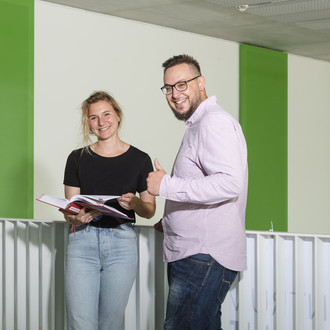 Zwei Studierende stehen vor einem Geländer, einer streckt den Daumen hoch.