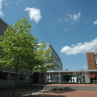Rektorat und Experimentierhalle: Rechts im Bild das Rektorat, ein graues Gebäude mit Fensterfronten, in der Mitte eine Treppe und links eine graue Gebäudefront mit einem Baum davor. Im Hintergrund links ein weiteres hohes Gebäude.