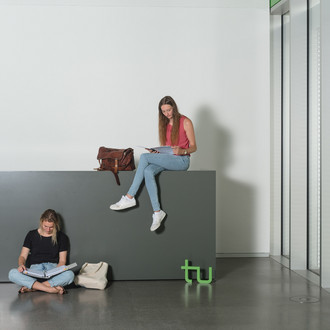 Eine Studierende sitzt auf einer Theke und liest, eine andere sitzt auf dem Boden und liest auch. Rechts im Bild ein großes Fenster.