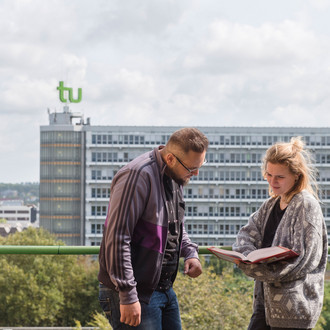 Ein Student und eine Studentin sehen gemeinsam in ein Buch, im Hintergrund ist der Mathetower in seiner vollen Länge zu sehen.
