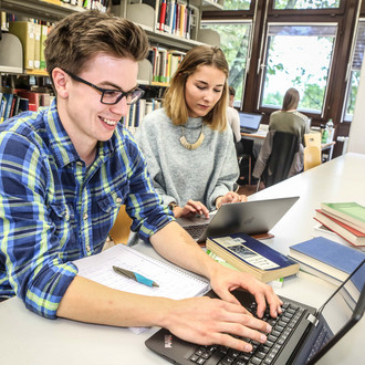Zwei Studierende sitzen in der Bibliothek und lernen am Laptop. 