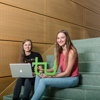 Zwei Studentinnen sitzen mit Laptop und TU-Logo auf einer Treppe.