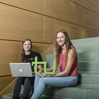 Zwei Studentinnen sitzen mit Laptop und TU-Logo auf einer Treppe.