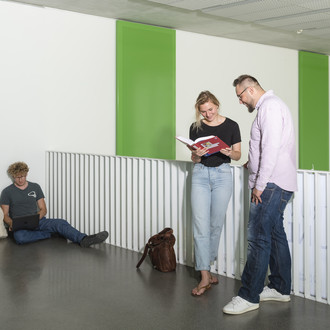 Studierende stehen vor einem Geländer und sehen in ein Buch, ein anderer sitzt auf dem Boden.