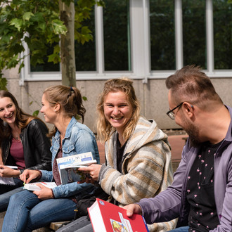 Vier Studierende sitzen auf einer Bank mit Büchern in der Hand und unterhalten sich.