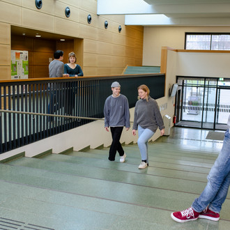 Studierende im Seminargebäude 1, im Vordergrund steht ein Student am Geländer und schaut auf zwei andere Studierende, die die Treppe hochgehen.