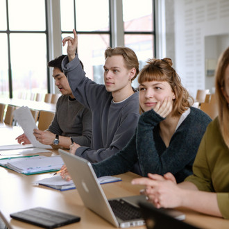 Studierende im Seminarraum sitzen an den Tischen. Eine Person meldet sich. 