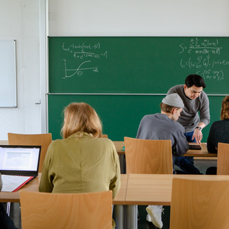 Vier Studierende im Seminarraum sitzen und arbeiten zusammen mit dem Dozierenden im intergrund.