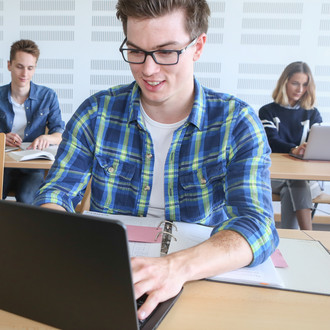 Student sitzt im Seminarraum am Laptop, im Hintergrund weitere Studierende